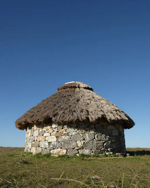 Round dry-stone herders hut with a thatched roof stands on a grassy plain under a clear deep blue sky