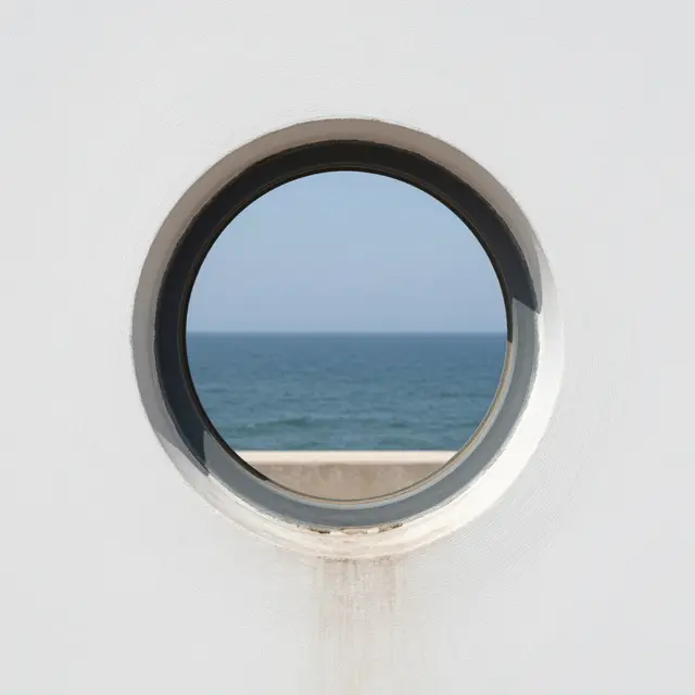 A round porthole window in a white wall frames a view of the calm blue sea under clear sky