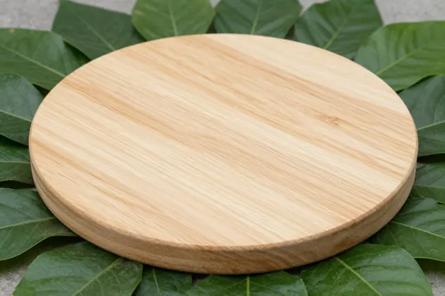 A smooth light wooden round display plinth rests surrounded by fresh green leaves on a gray surface