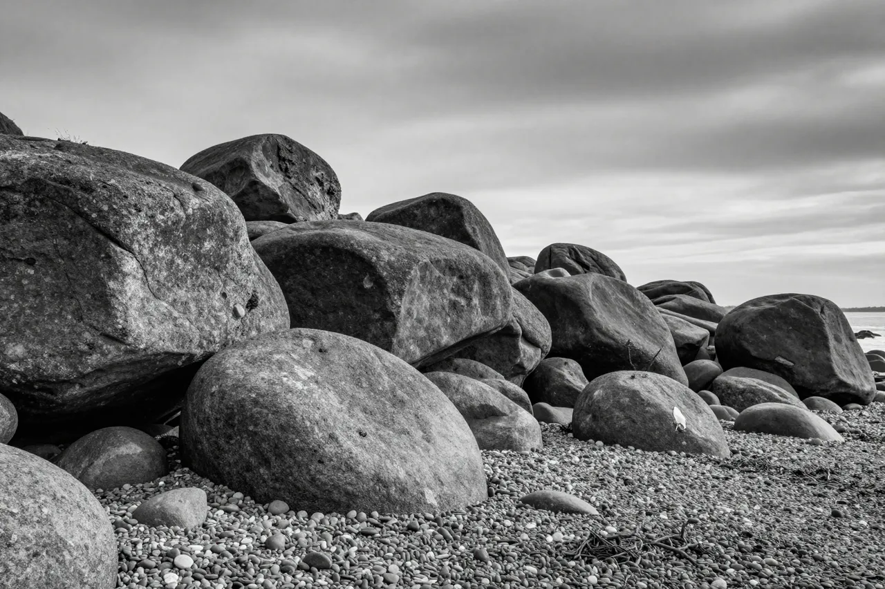 Black and white view of large weathered basalt boulders resting on a pebbled shore under an overcast heavy sky