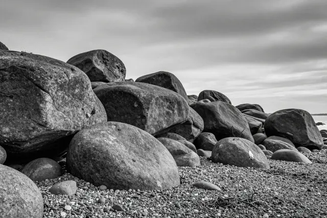 Black and white view of large weathered basalt boulders resting on a pebbled shore under an overcast heavy sky