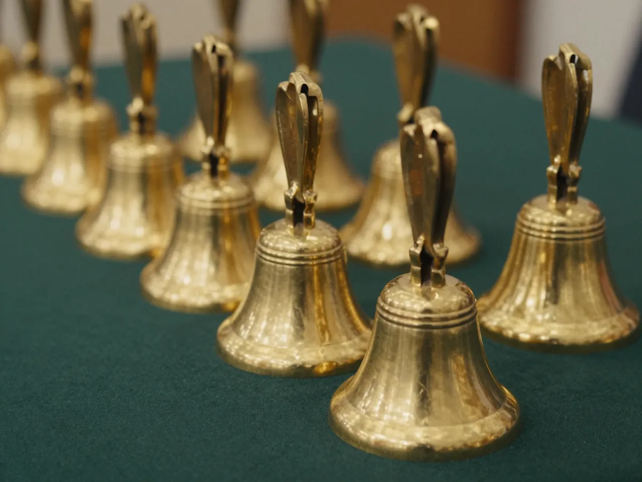 A diagonal row of polished brass handbells rests on a dark green fabric surface with warm soft lighting.