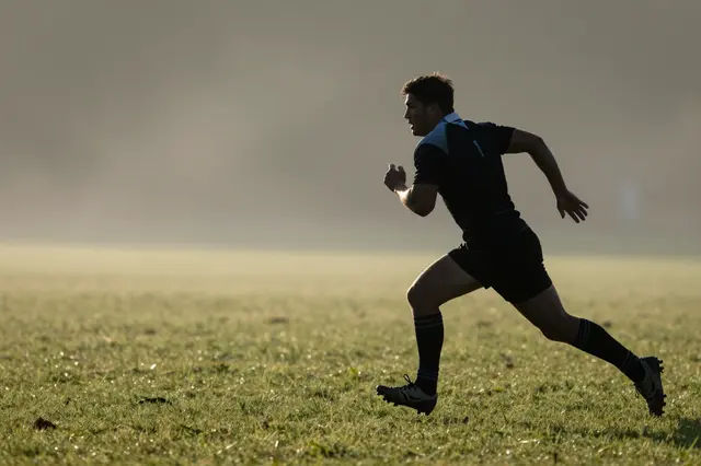 An adult rugby player runs across a dewy grass field in soft dawn mist