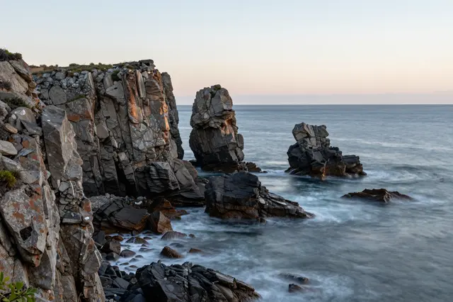 Layered sea stacks and rocky cliffs rise from a calm ocean under a pale dawn sky.