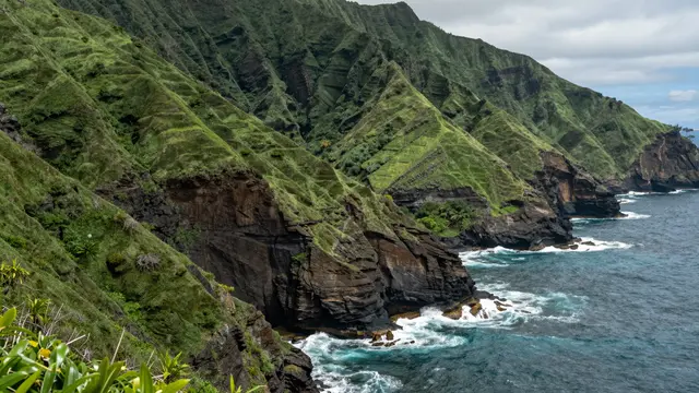Lush ridged green coastal cliffs meet turquoise ocean waves under an overcast sky
