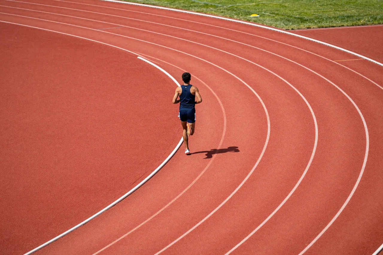 A runner in dark athletic wear rounds the curve of a red running track viewed from above, casting a sunlit shadow.