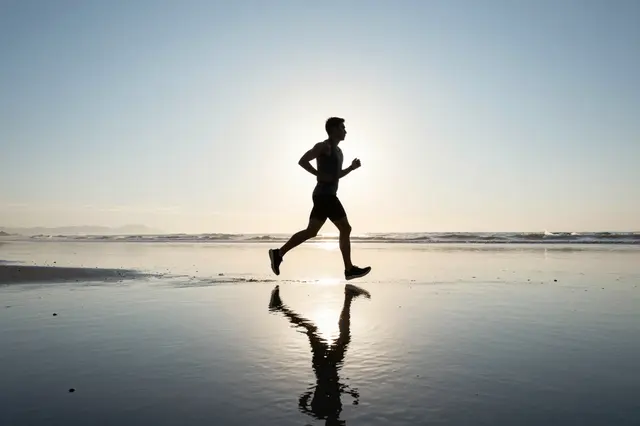 Silhouetted adult runner jogs across wet low-tide beach with his reflection visible at sunrise