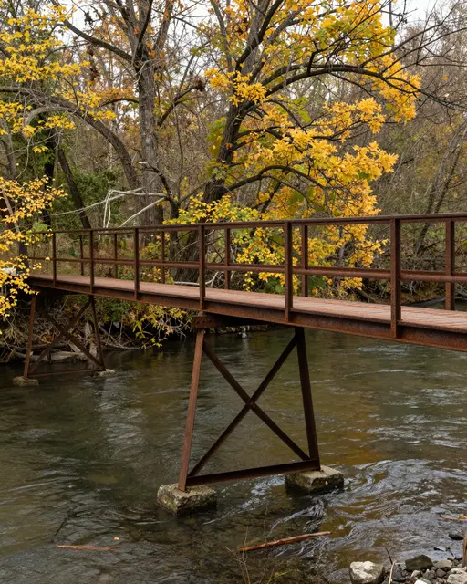 A weathered rusted iron footbridge spans a quiet river beneath yellow autumn trees