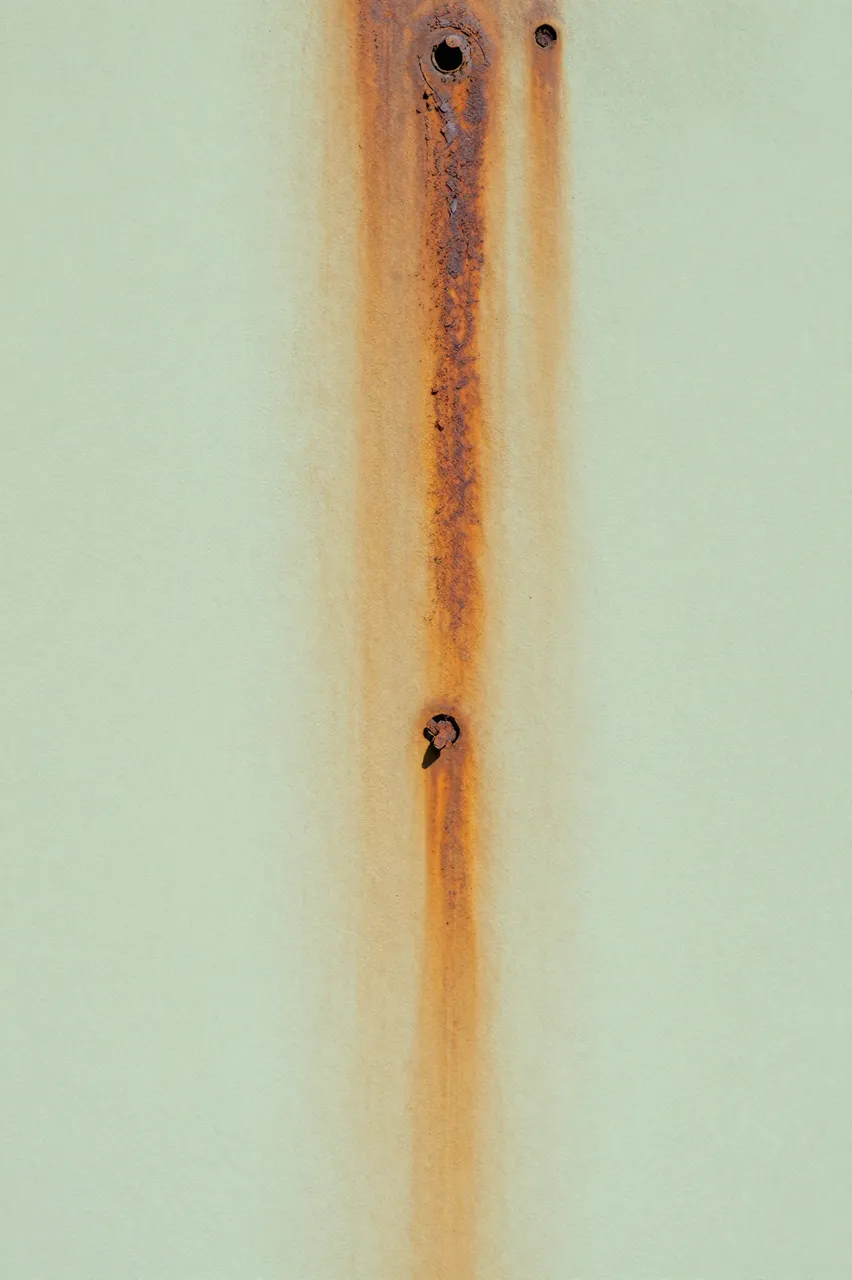 Vertical rust streaks run down a pale green painted wall around old nail holes.