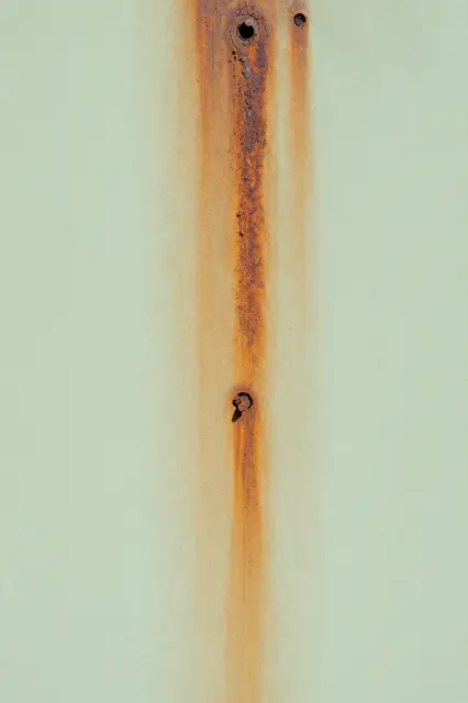 Vertical rust streaks run down a pale green painted wall around old nail holes.