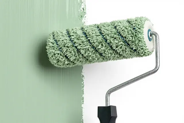 Paint roller loaded with sage green paint rests against a wall with a fresh painted edge next to a white surface
