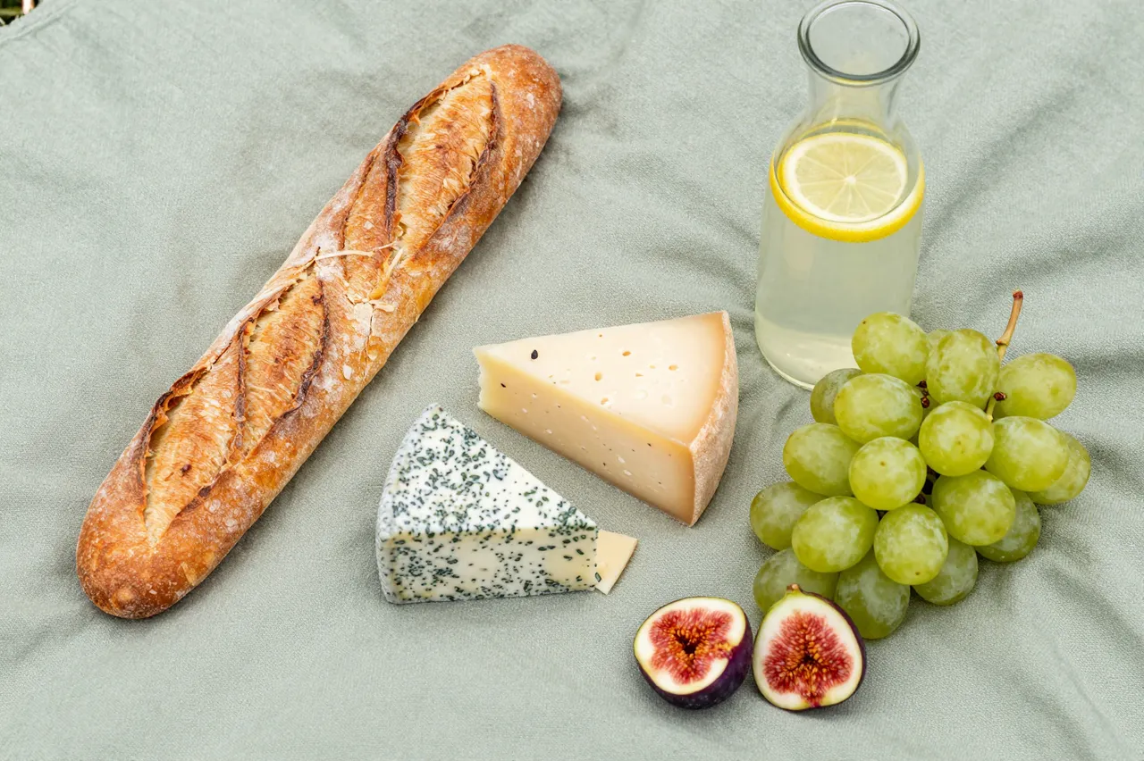 Flat lay of a picnic with baguette, cheeses, green grapes, sliced figs and lemonade on a sage linen blanket