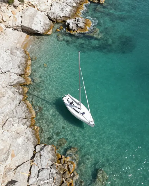 Aerial top-down view of a white sailboat anchored in clear turquoise water beside pale rocky shoreline.