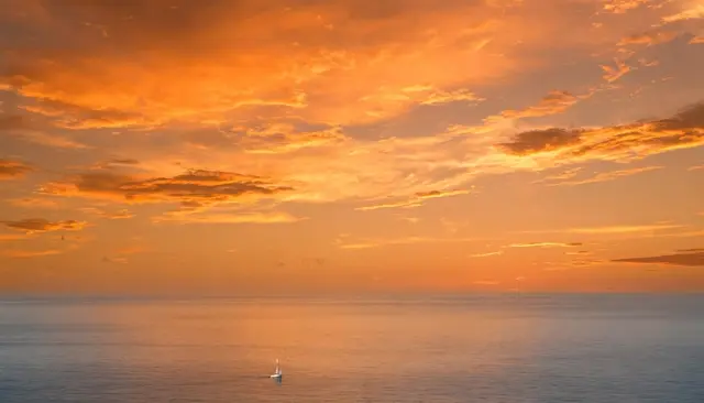 A small white sailboat floats on calm glassy sea beneath a vast vibrant orange sunset sky.