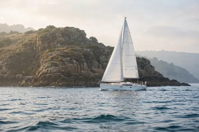 White sailboat with plain sails glides on calm water past a rocky coastal headland in soft morning light