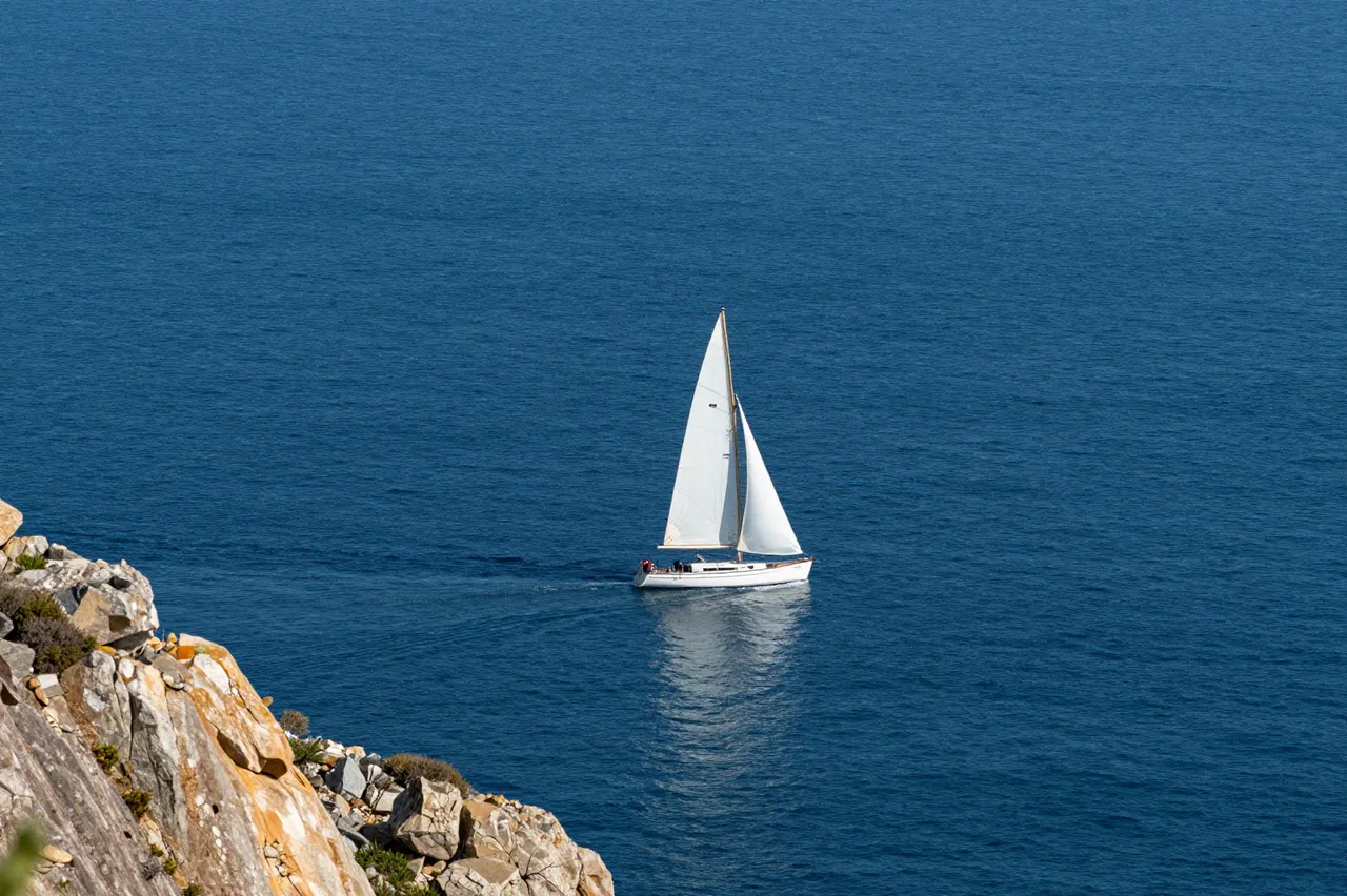 High angle view of a lone white sailboat floating on deep blue ocean next to a rocky cliff edge