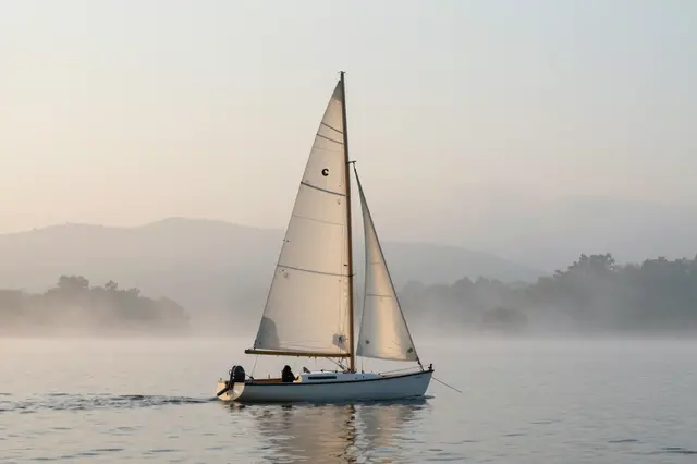A small sailboat floats on calm misty lake waters beneath pale dawn skies with distant foggy hills.