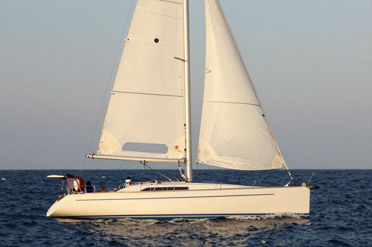 A white sailboat with unfurled sails travels across dark open ocean under a pale clear sky during golden hour