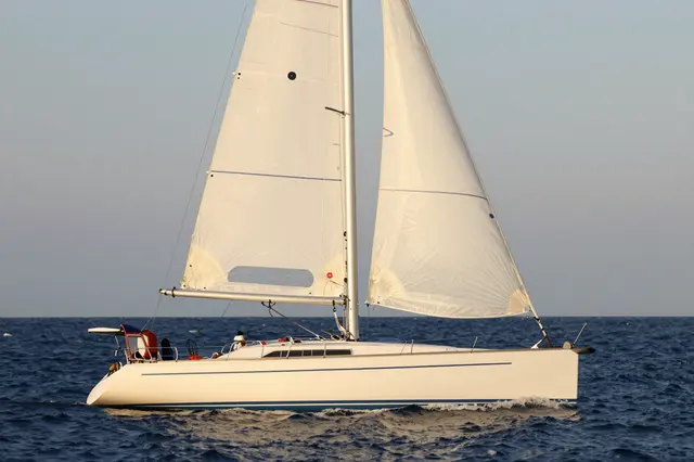 A white sailboat with unfurled sails travels across dark open ocean under a pale clear sky during golden hour