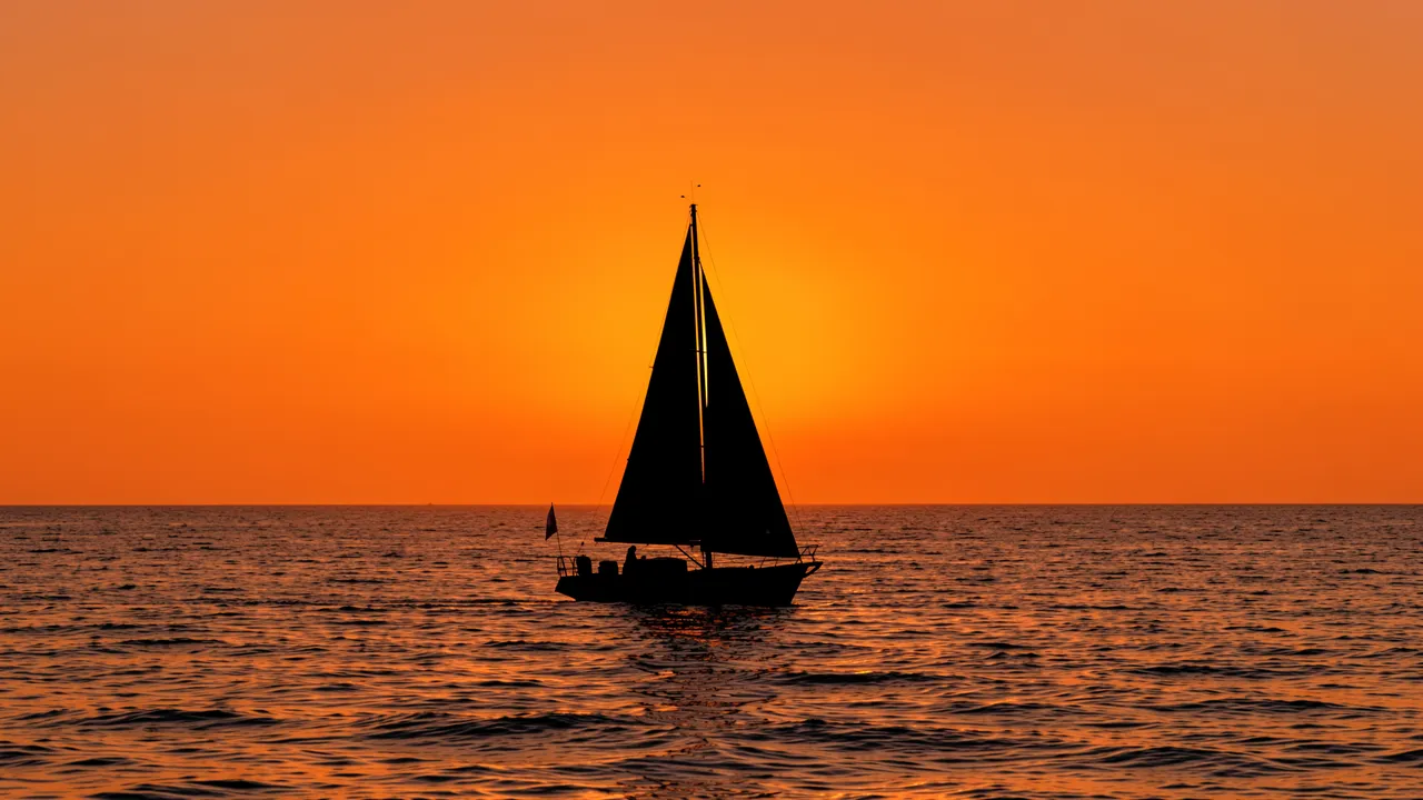 Black silhouette of a small sailboat on rippled sea beneath a bright tangerine sunset sky