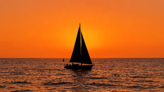 Black silhouette of a small sailboat on rippled sea beneath a bright tangerine sunset sky