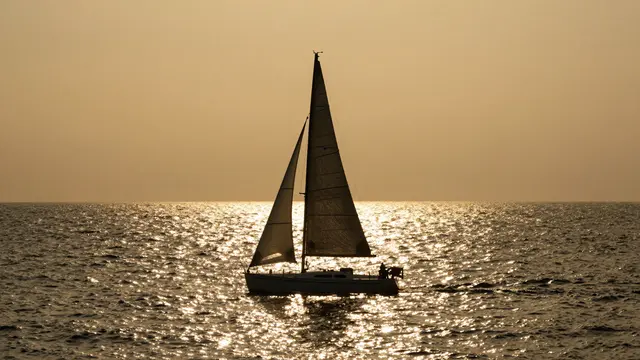 Silhouetted sailboat floats on glittering golden sunlit sea at sunset