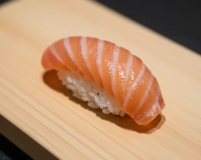 A single salmon nigiri rests centered on a light wooden sushi board against a dark background