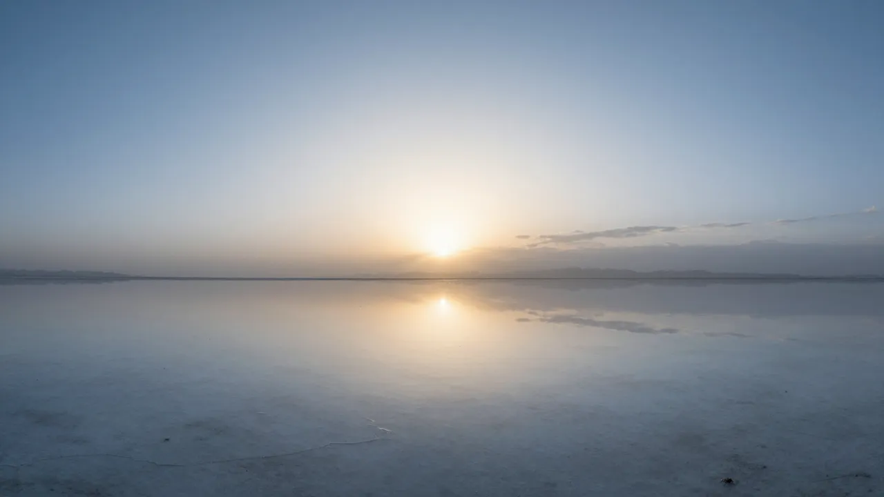 The rising sun casts a sharp mirror reflection across still, flooded salt flats under a pale dawn sky.