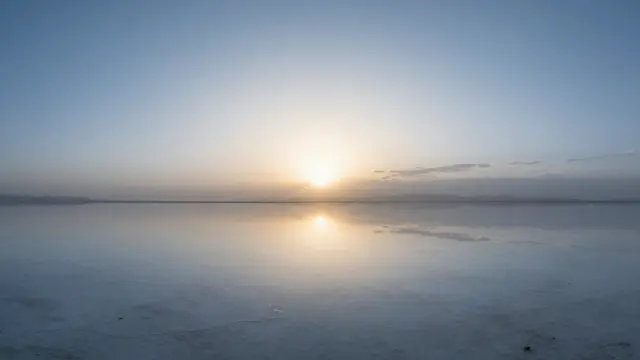 The rising sun casts a sharp mirror reflection across still, flooded salt flats under a pale dawn sky.