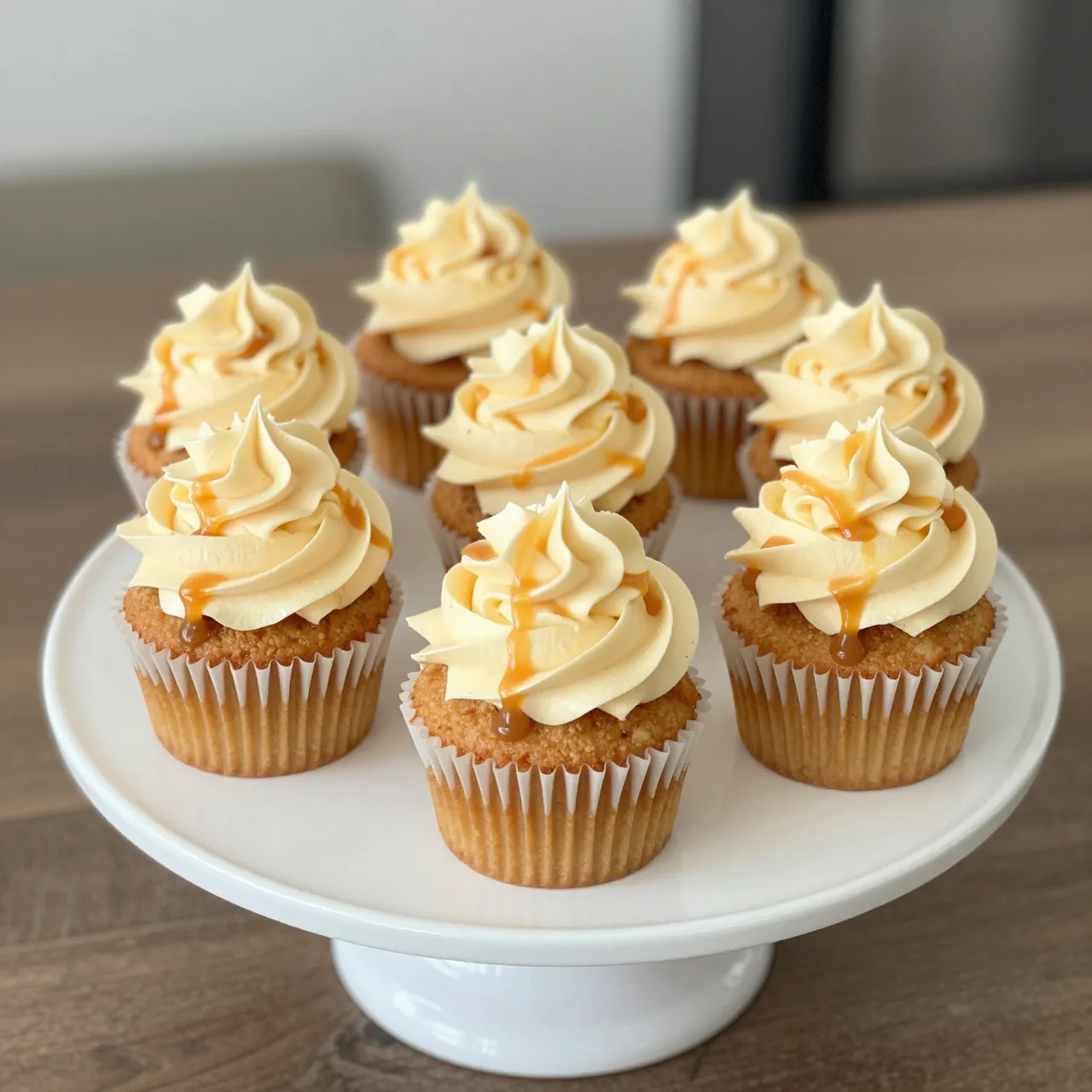 Eight salted caramel cupcakes with tall frosting swirls and caramel drizzle arranged on a white cake stand.