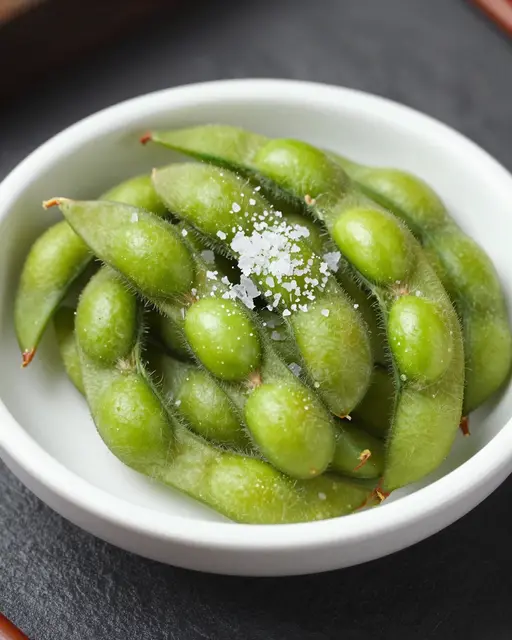 A close-up view of steamed edamame pods topped with flaky sea salt in a small white bowl