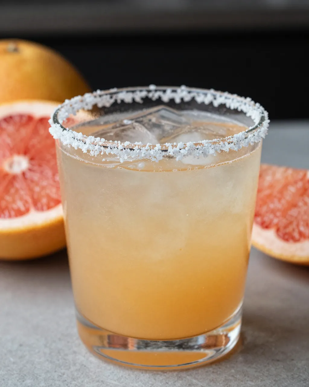 A paloma cocktail with a salted rim sits surrounded by fresh grapefruit slices on a light countertop