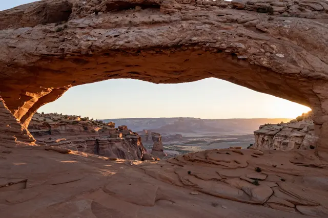 A natural sandstone arch frames a distant desert mesa landscape at golden sunrise