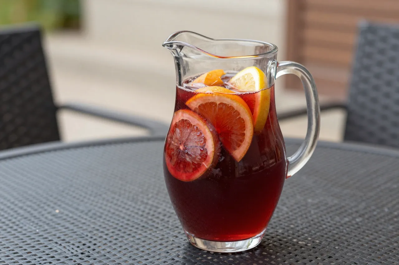 A glass pitcher of red sangria with sliced citrus fruit rests on an outdoor patio table.