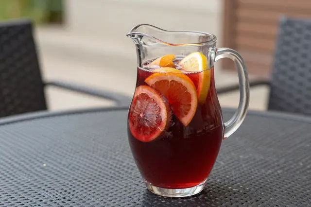 A glass pitcher of red sangria with sliced citrus fruit rests on an outdoor patio table.