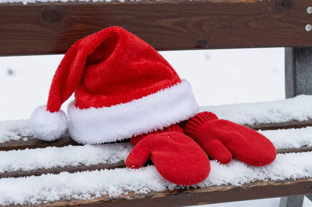 A fuzzy red Santa hat with white trim rests beside matching red mittens on a snow-dusted wooden park bench.