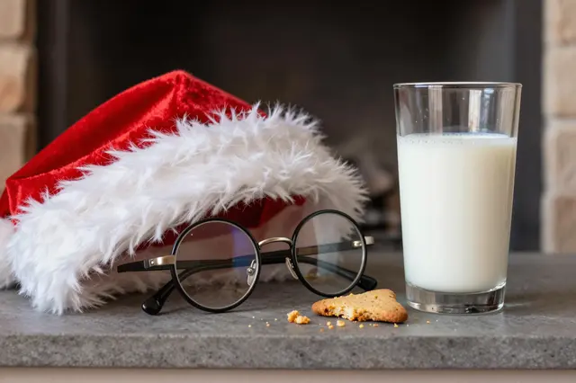 Santa hat, round spectacles, half-full milk glass and partially eaten cookie with crumbs rest on a stone fireplace hearth