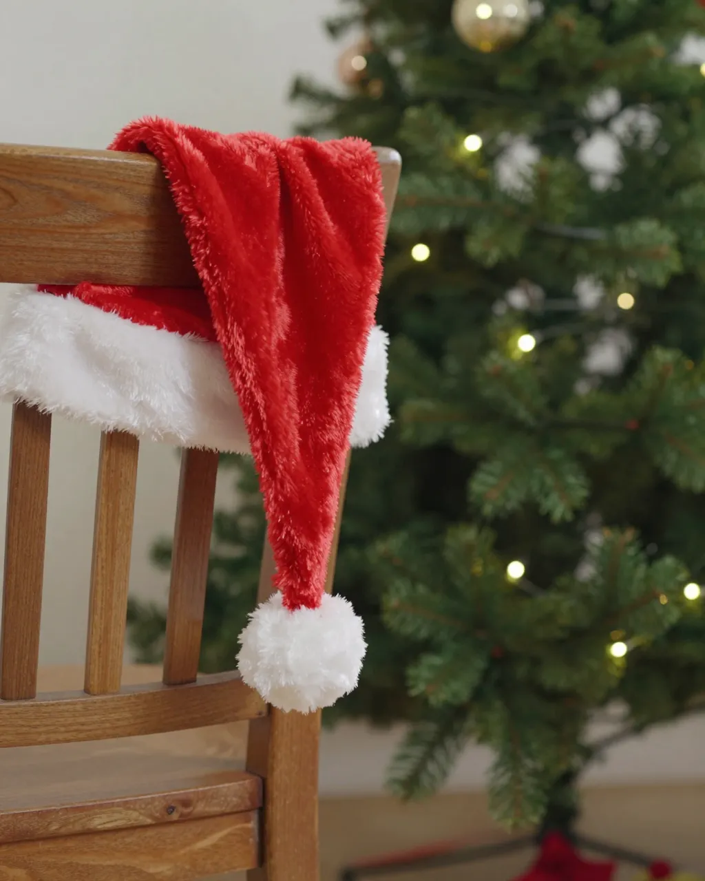 Plush red Santa hat with white fur trim hangs on the back of a wooden chair beside a glowing lit Christmas tree.