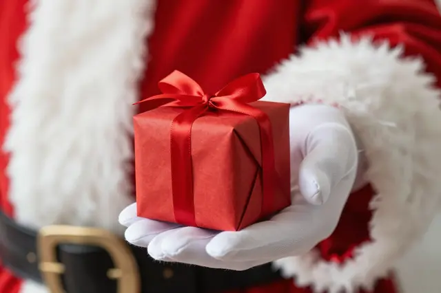 Close-up of Santa's white gloved hand holding out a small red wrapped gift with a bow