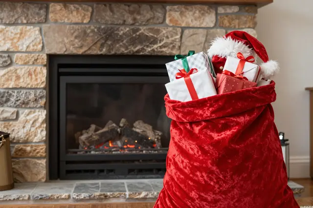 Red velvet Santa sack overflowing with wrapped gifts sits next to a lit stone fireplace