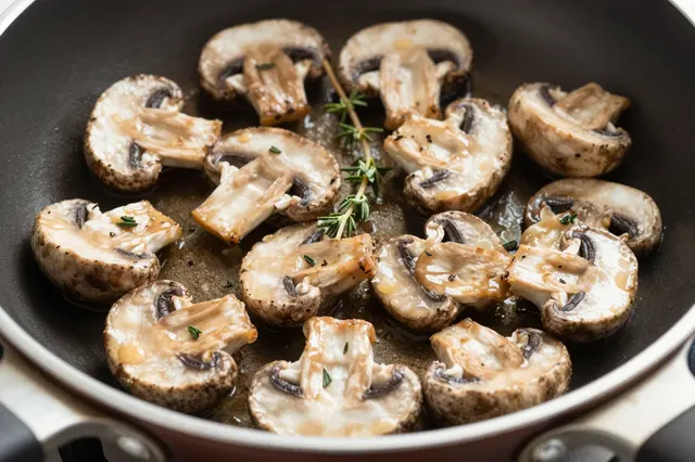 Sliced sauteed button mushrooms with fresh thyme sit glistening in a nonstick frying pan