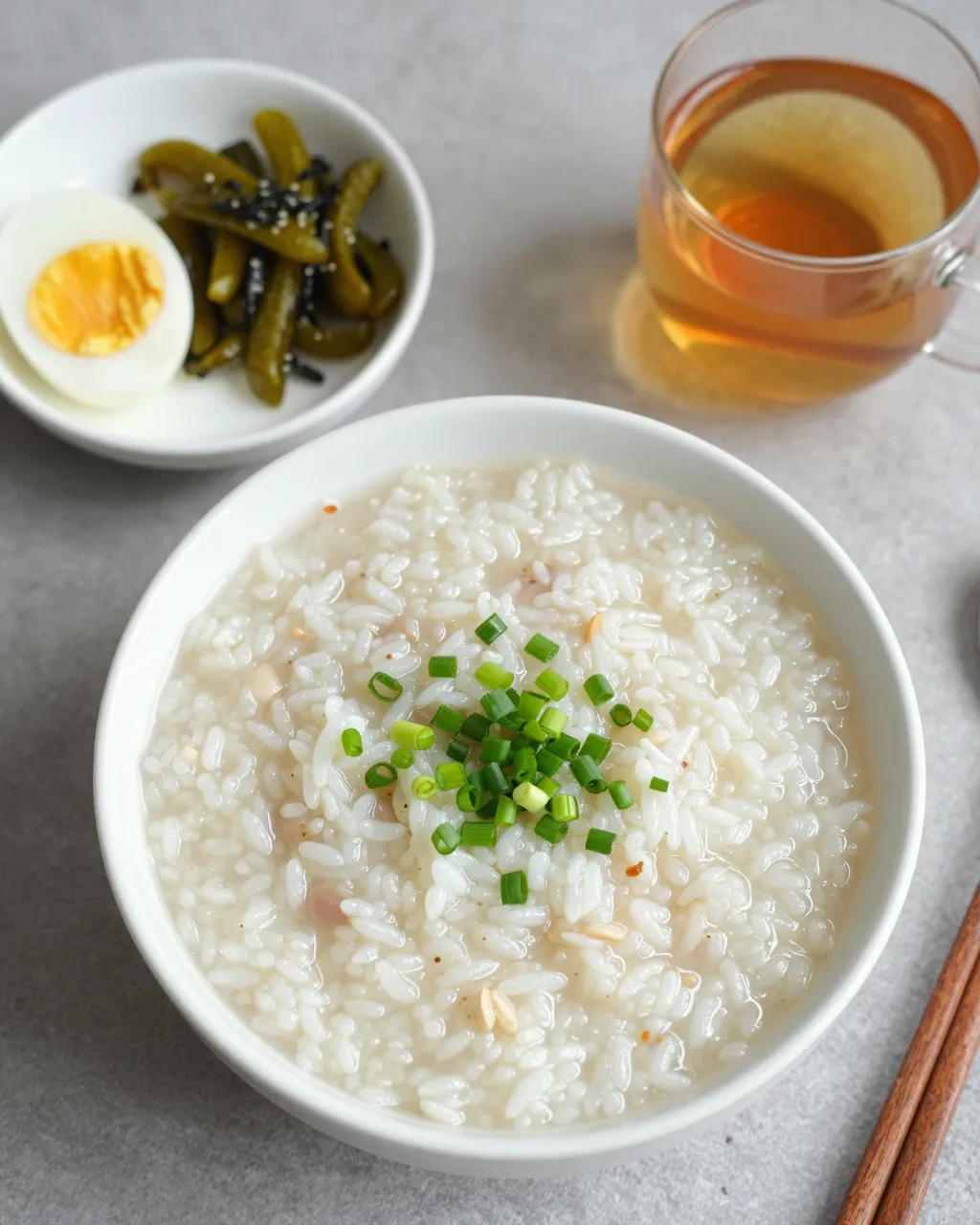 Flat lay of congee breakfast with toppings, pickles, boiled egg and tea on a gray countertop