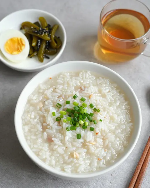 Flat lay of congee breakfast with toppings, pickles, boiled egg and tea on a gray countertop