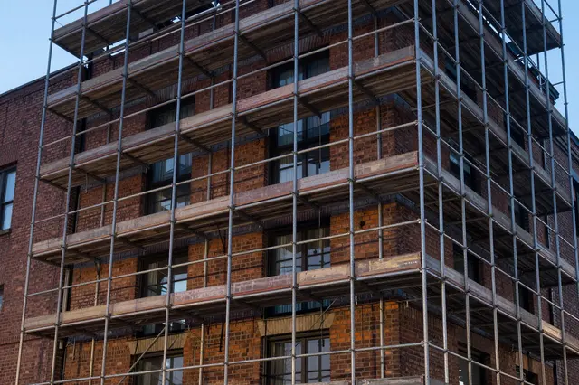 Metal and wooden scaffolding wraps the facade of a red brick multi-story building under a pale dusk sky