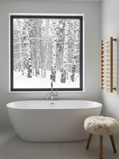 Freestanding white bathtub sits below a window showing snow-covered birch trees outside