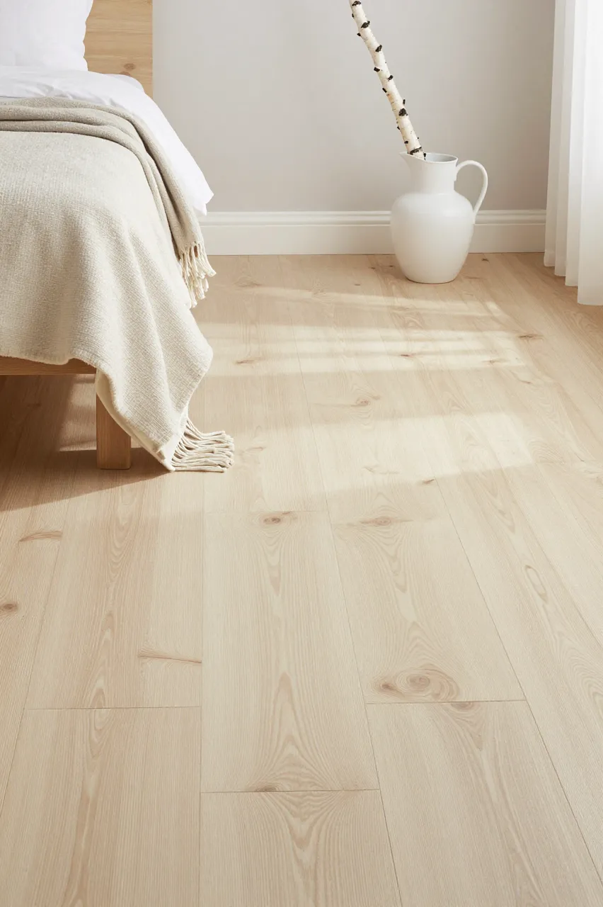 Sunlit pale pine bedroom floor with a draped wool blanket, bed edge, and birch branch in a white jug