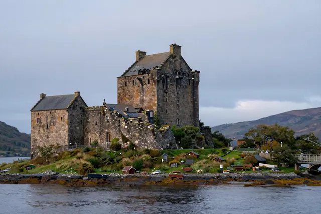 Stone castle ruin sits on a grassy islet at the edge of a calm Scottish loch under an overcast dusk sky