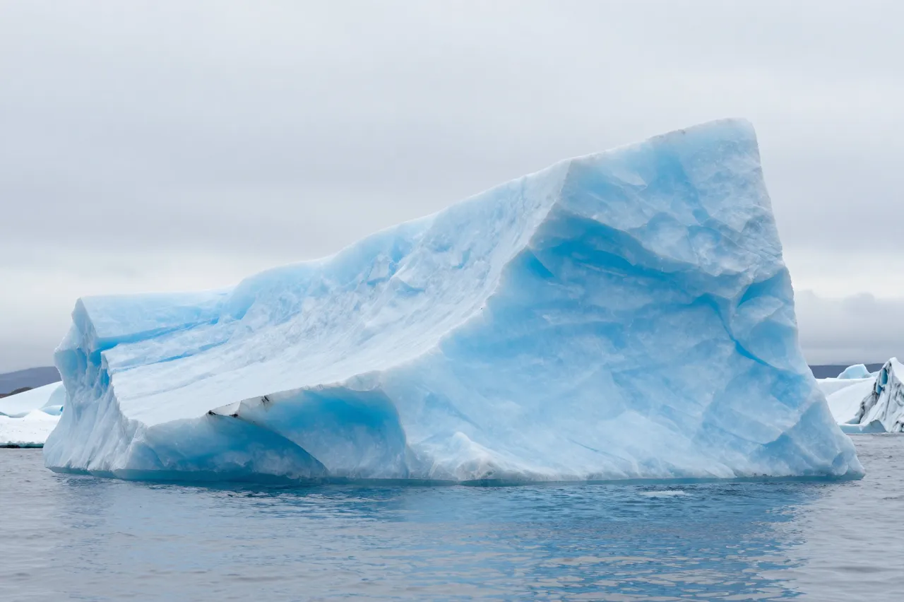 A large smooth blue iceberg floats in calm cold water under an overcast sky
