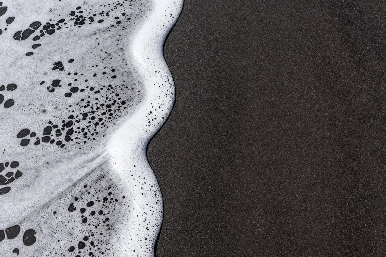 Top-down view of bubbly white sea foam resting along the edge of dark volcanic sand with empty copy space to the right.