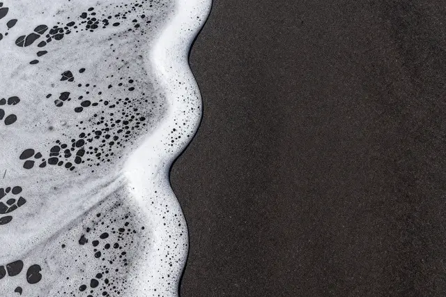 Top-down view of bubbly white sea foam resting along the edge of dark volcanic sand with empty copy space to the right.